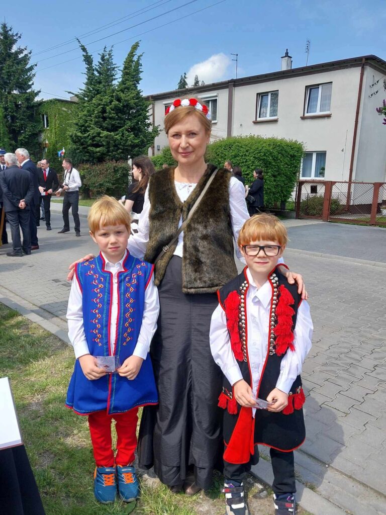 Kobieta w białej bluzce oraz brązowej kamizelce i w biało-czerwonej opasce na głowie pozuje do zdjęcia z dwoma chłopcami w strojach ludowych