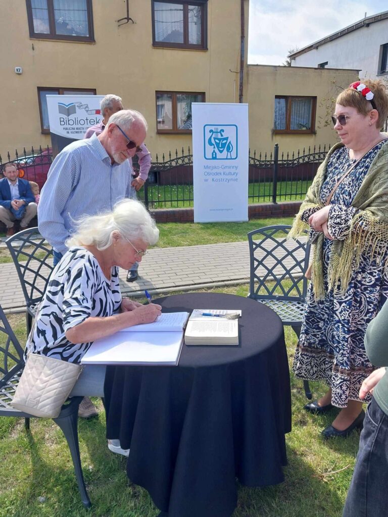 Kobieta przepisuje tekst do dużej księgi ułożonej na stole w plenerze, obok mężczyzna oraz kobieta