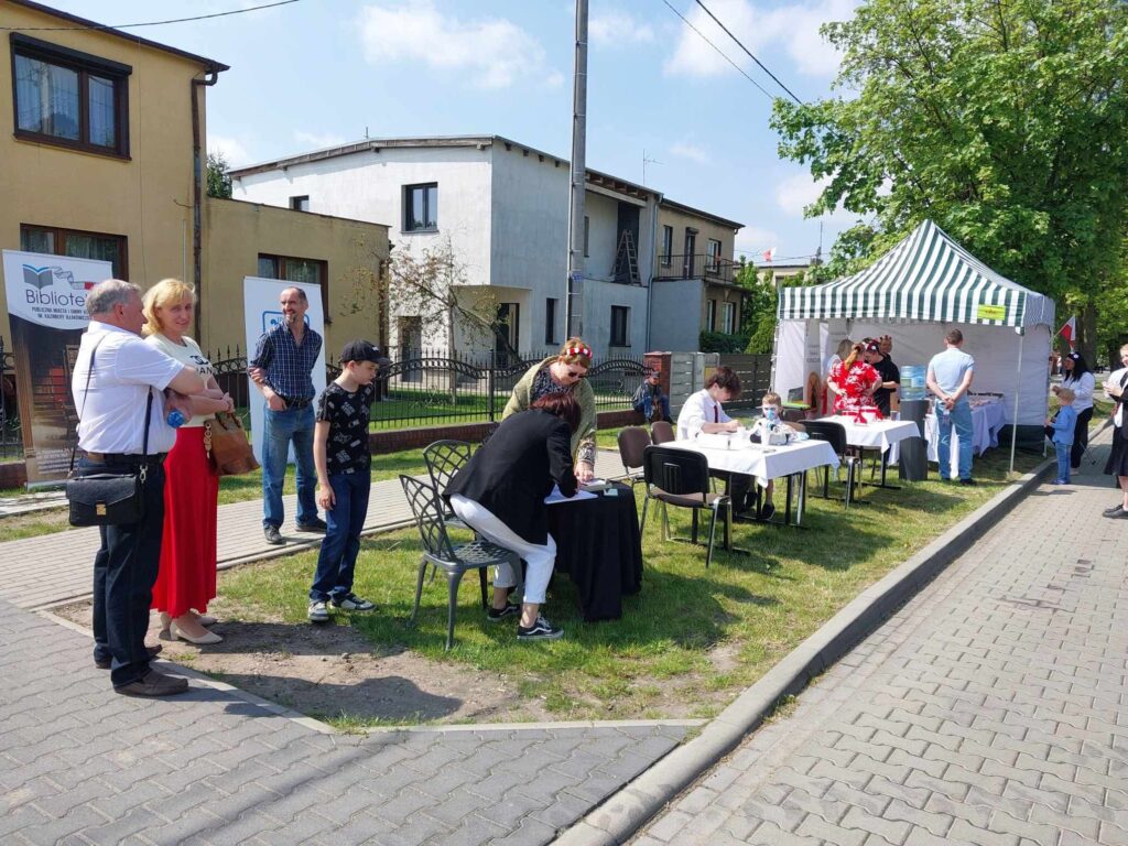 Przy stolikach ustawionych na trawniku przy drodze siedzą i stoją ludzie, w tle duży namiot imprezowy