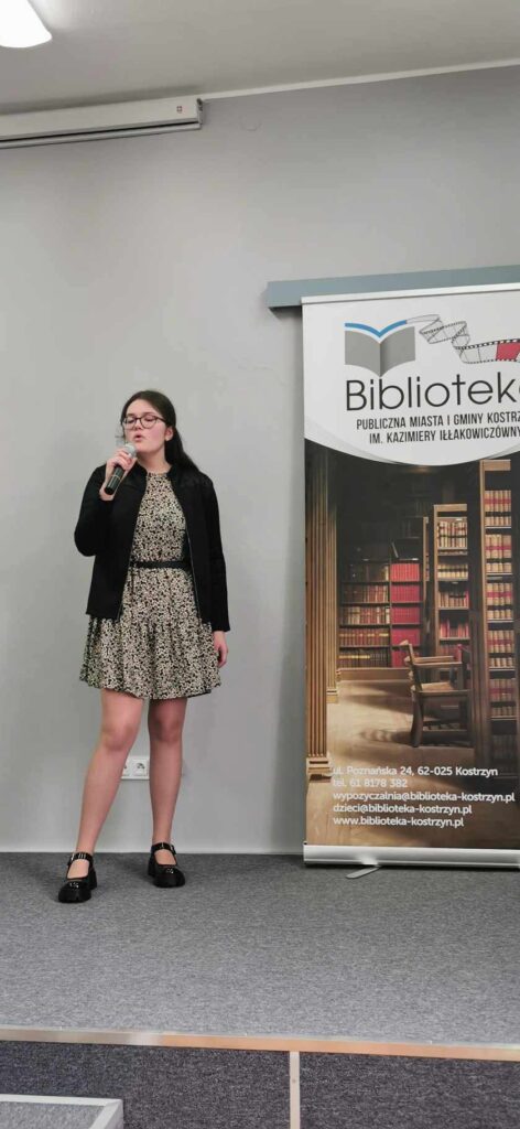 Dziewczynka w czarnej bluzie i sukience śpiewa piosenkę na scenie w tle baner biblioteki