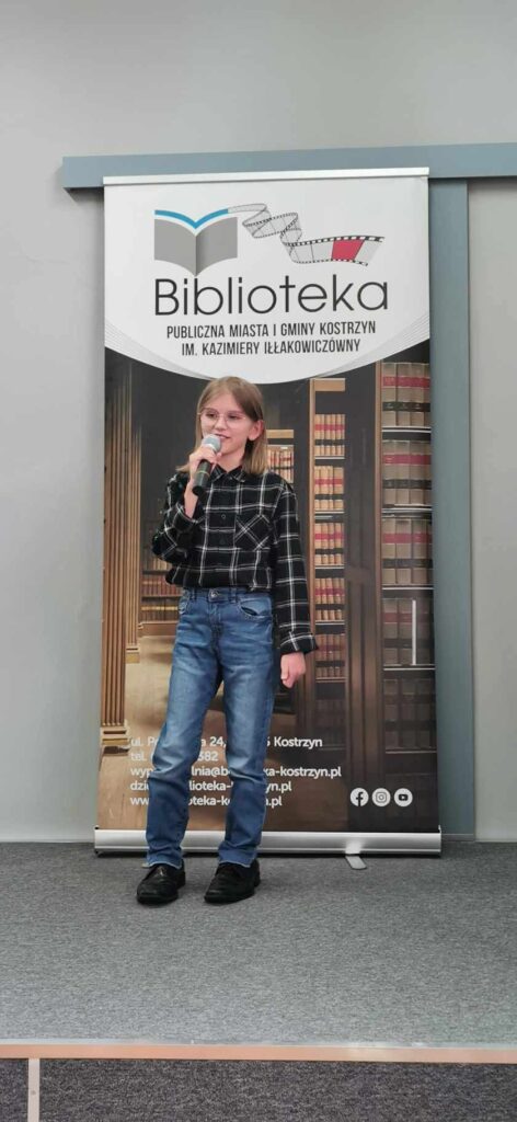Dziewczynka w koszuli w kratkę recytuje wiersz na scenie w tle baner biblioteki
