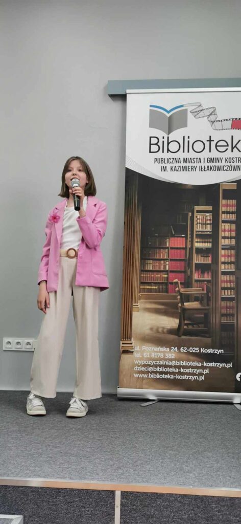 Dziewczynka w różowej bluzie recytuje wiersz na scenie w tle baner biblioteki