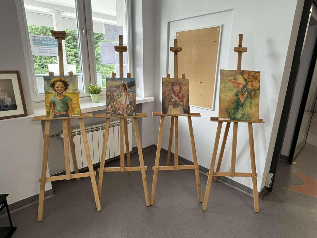 Na czterech sztalugach w holu biblioteki stoją obrazy przedstawiające dzieci.
