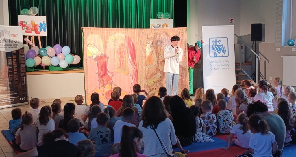 Zdjęcie przedstawia salę, w której odbywa się spektakl dla dzieci. Na tle kolorowej scenografii stoi aktor, dzieci siedzą na podłodze przed nim.