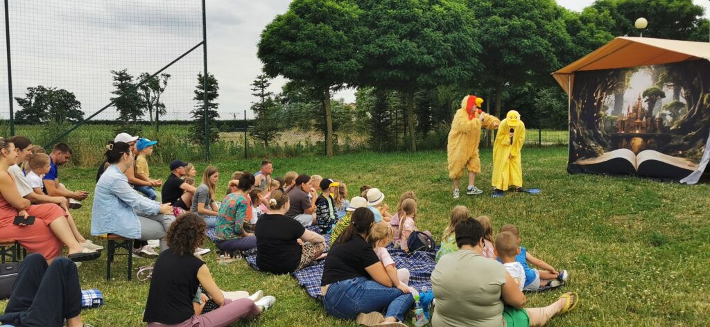 Publiczność siedzi na kocach i ławkach oglądając spektakl w Iwnie