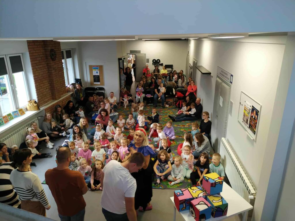 Hol biblioteki widziany ze schodów na piętro - na dywanach siedzą dzieci wraz z opiekunami.