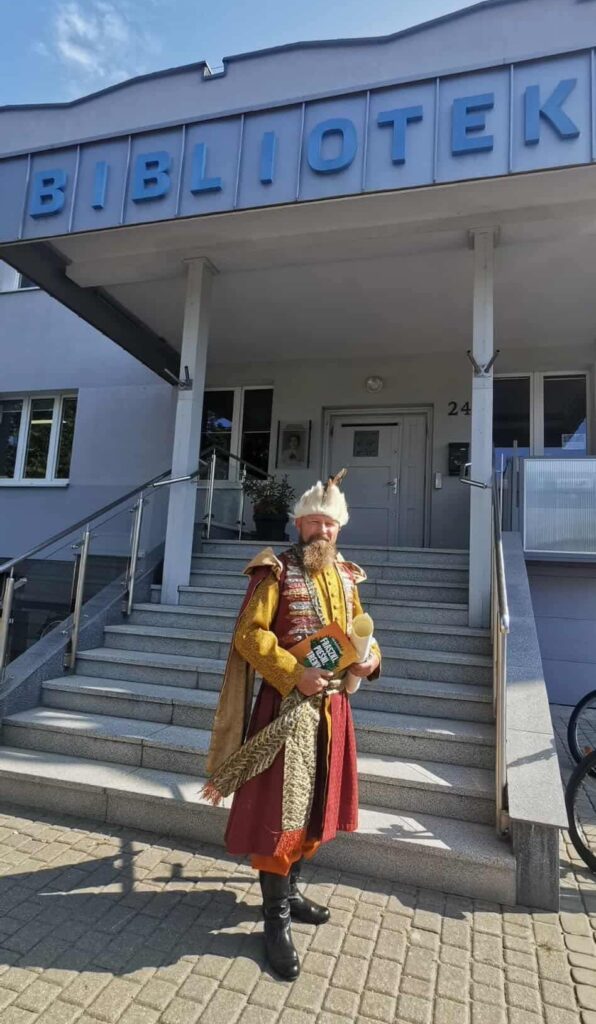 Przed budynkiem biblioteki stoi mężczyzna z brodą, w stroju szlacheckim.
