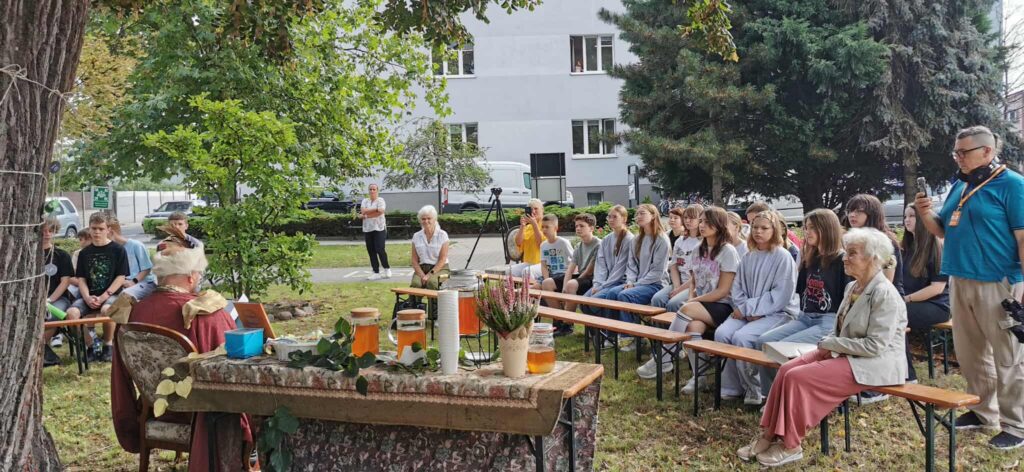 Na skwerze pod lipą Grupa młodzieży siedzi na ławkach. 