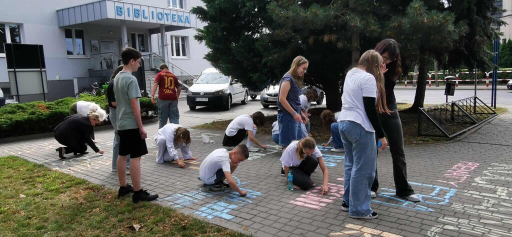 Grupka młodzieży pisze kredą  na chodniku przed biblioteką.