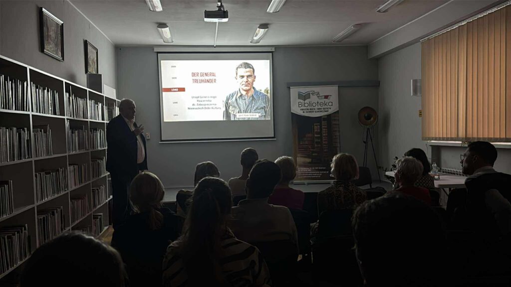 Na zdjęciu przyciemniona sala. Mężczyzna stojący obok ekranu wyświetla prezentację.