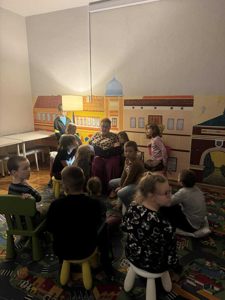 Bibliotekarka siedząca w fotelu czyta zgromadzonym wokół dzieciom. Z tyłu lampa rzuca delikatne światło.