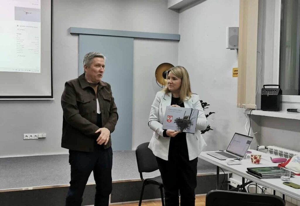 Na zdjęciu stoją: kobieta trzymająca w rękach album oraz mężczyzna w brązowej kurtce.