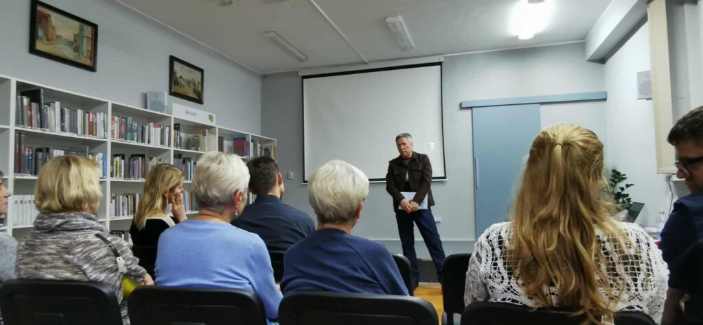 W sali z ekranem i rzutnikiem mężczyzna stoi przed widzami siedzącymi na krzesłach.