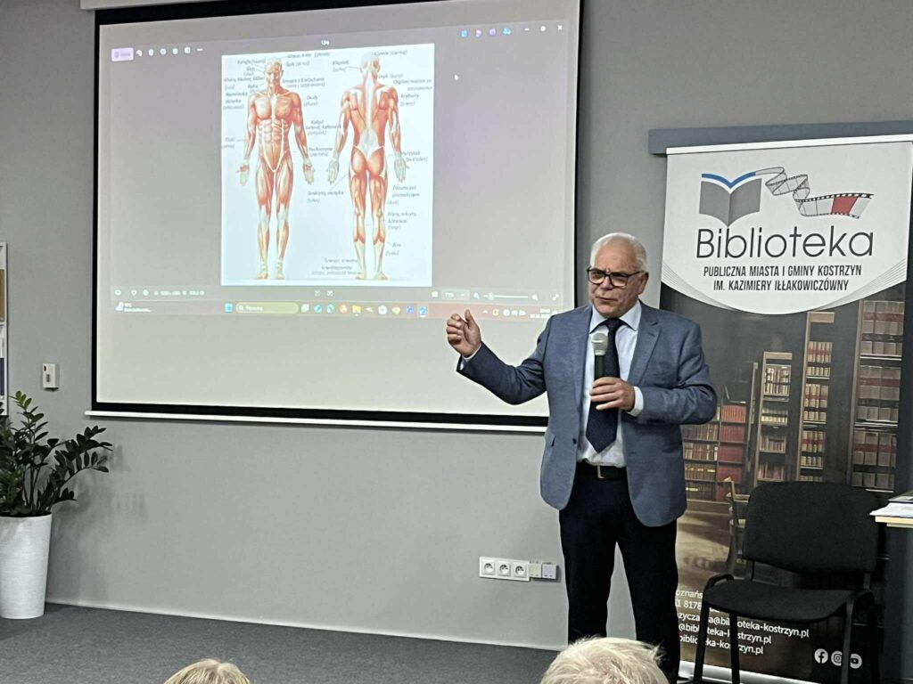 Mężczyzna w szarej marynarce prezentuje slajd na ekranie ściennym. Po prawej stronie rollup biblioteki.