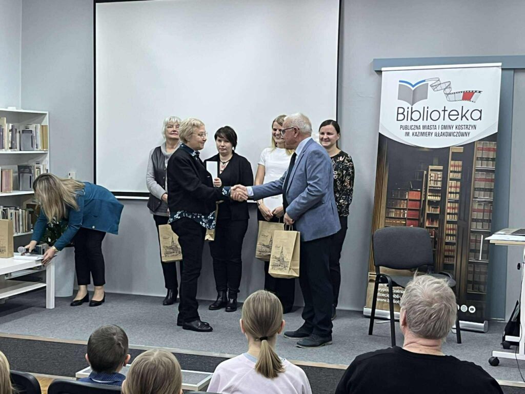 Na podeście w czytelni odbywa się wręczanie nagród. Mężczyzna gratuluje kobiecie w czarnym żakiecie.