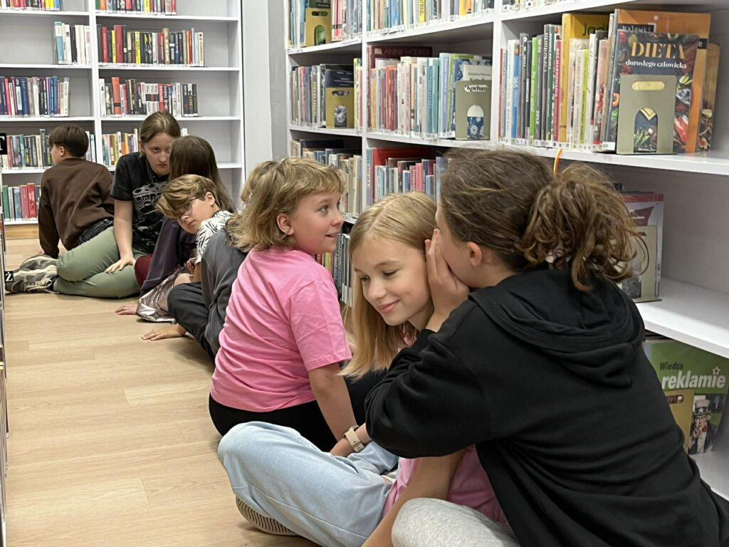 Na podłodze między regałami z książkami dzieci siedzące w rzędzie szepczą sobie do ucha.