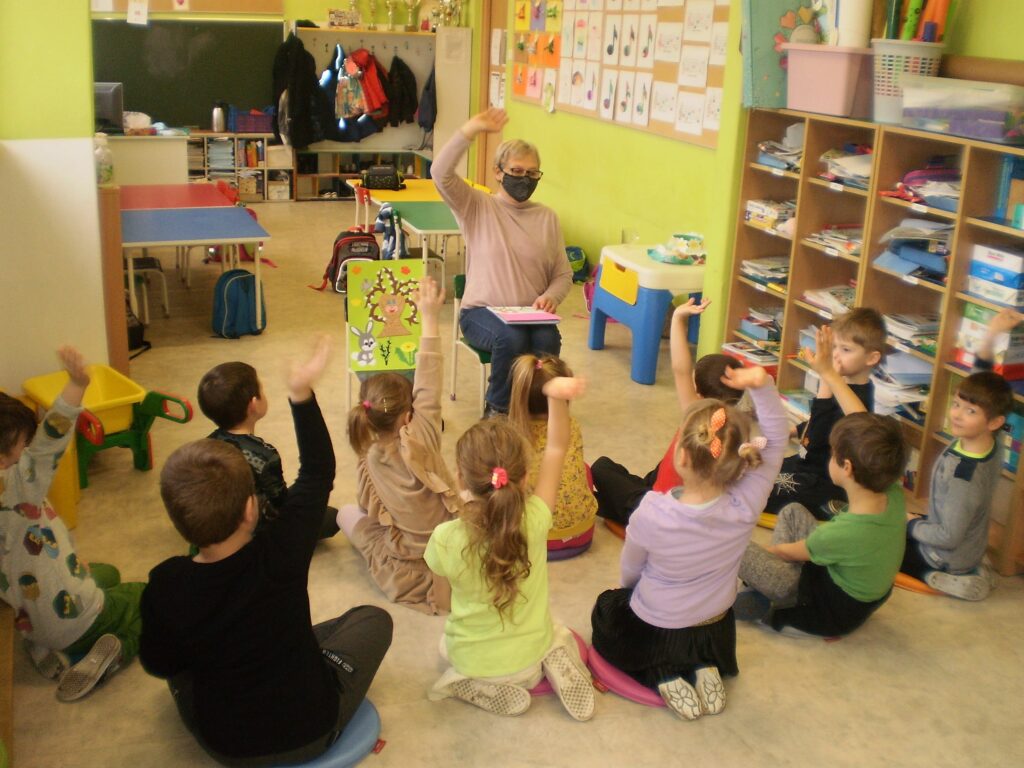 Nauczyciel siedzi na krześle, obok na sztaludze plakat wiosenny, dzieci siedzą na dywanie i zgłaszają się do odpowiedzi