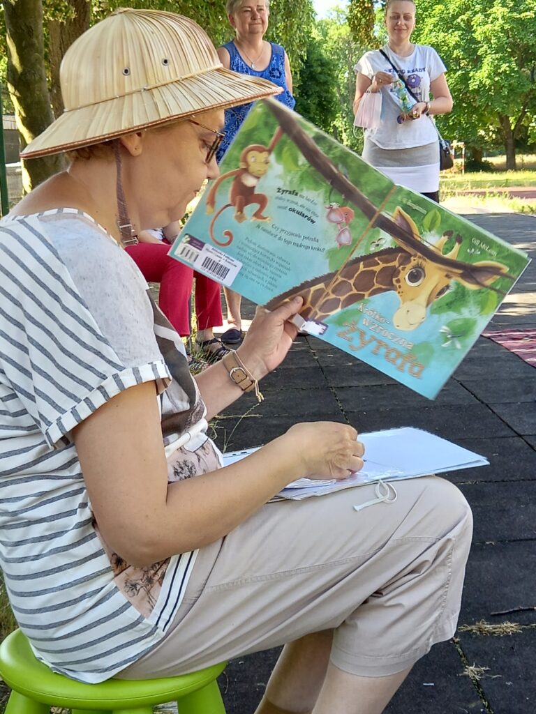 Bibliotekarka w cieniu drzewa czyta dzieciom książkę o żyrafie.