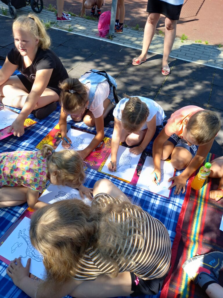Grupa dzieci zajęta rysowaniem na kocu rozłożonym pod drzewem.