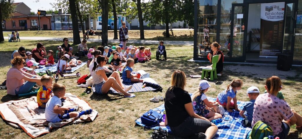 W parku na trawie na rozłożonych kocach siedzą dzieci. W tle Stacja Książki.