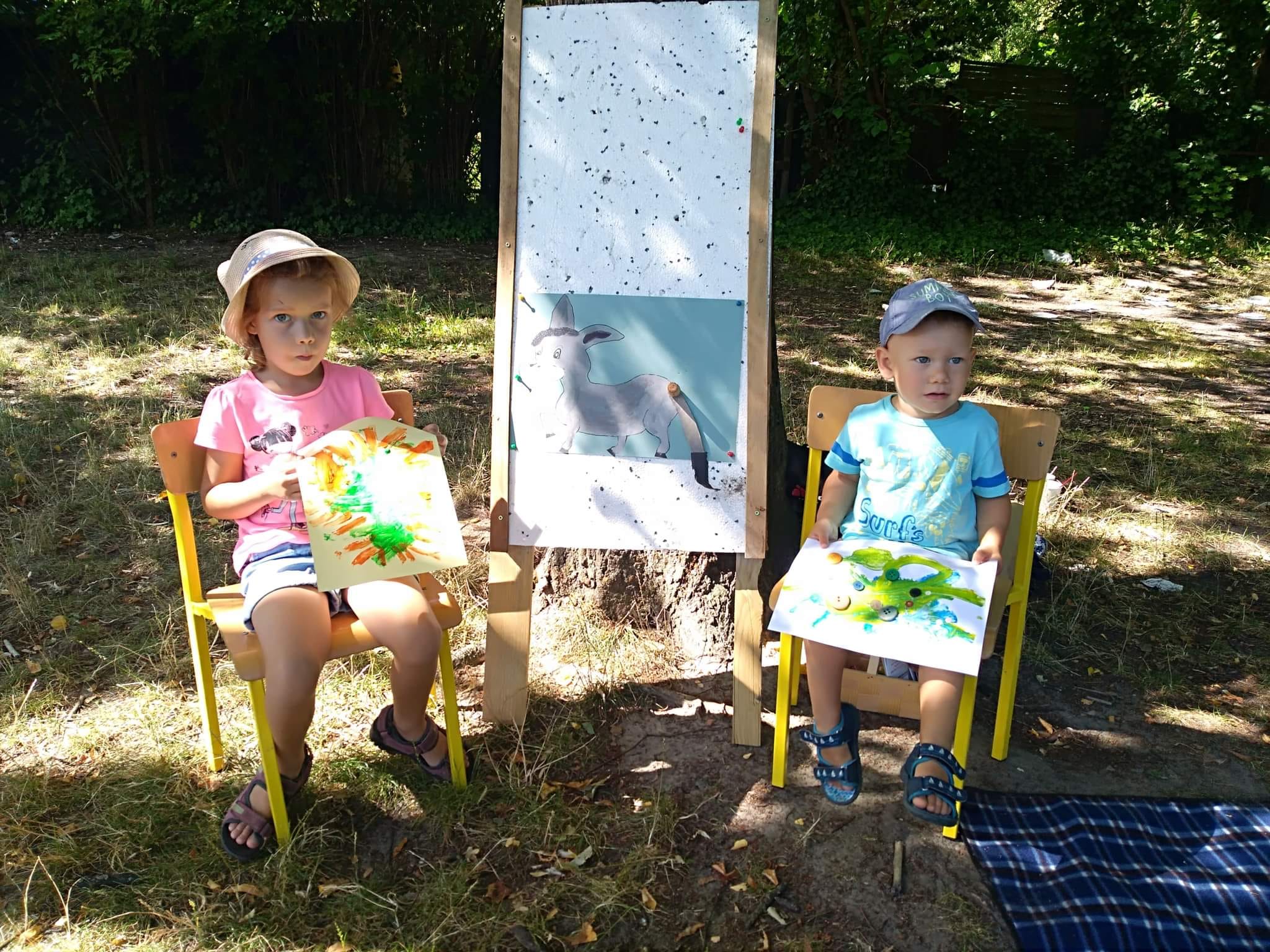 Dziewczynka oraz mały chłopiec siedzą na krzesełkach po bokach tablicy z narysowanym osiołkiem. Dzieci w rękach trzymają swoje rysunki.