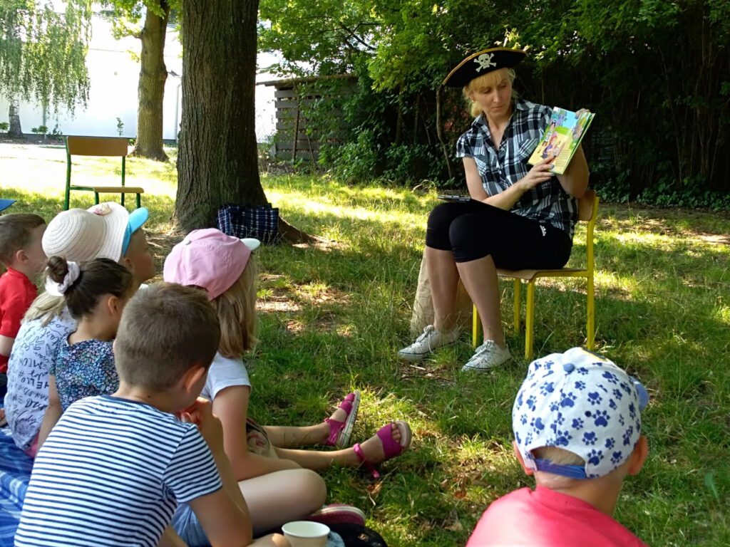 W cieniu drzew bibliotekarka w pirackiej czapce czyta dzieciom bajkę.