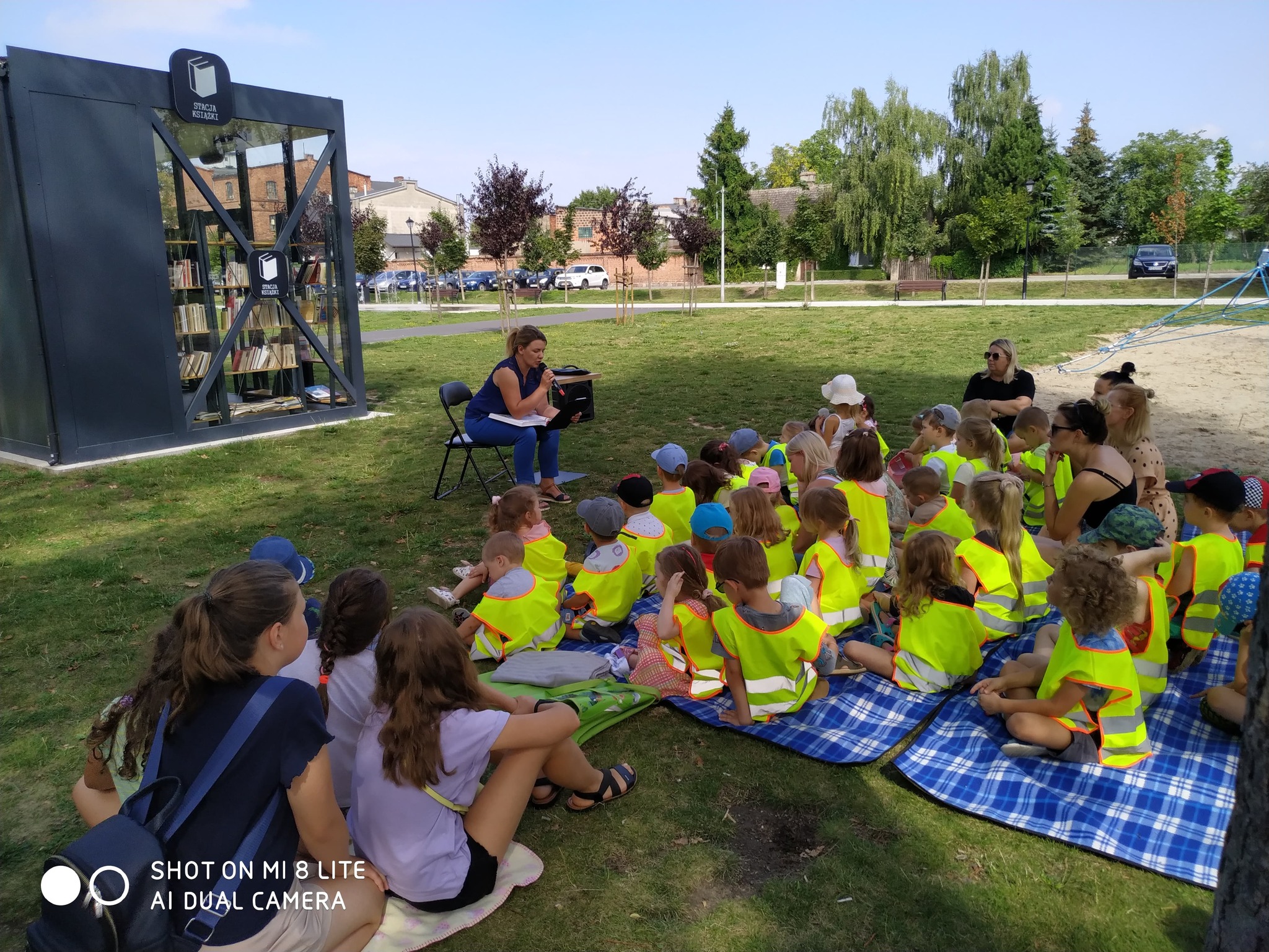 W Parku Miejskim Grupa przedszkolaków siedzi na trawie słuchając opowiadania czytanego przez panią dyrektor biblioteki. W tle Stacja Książki.