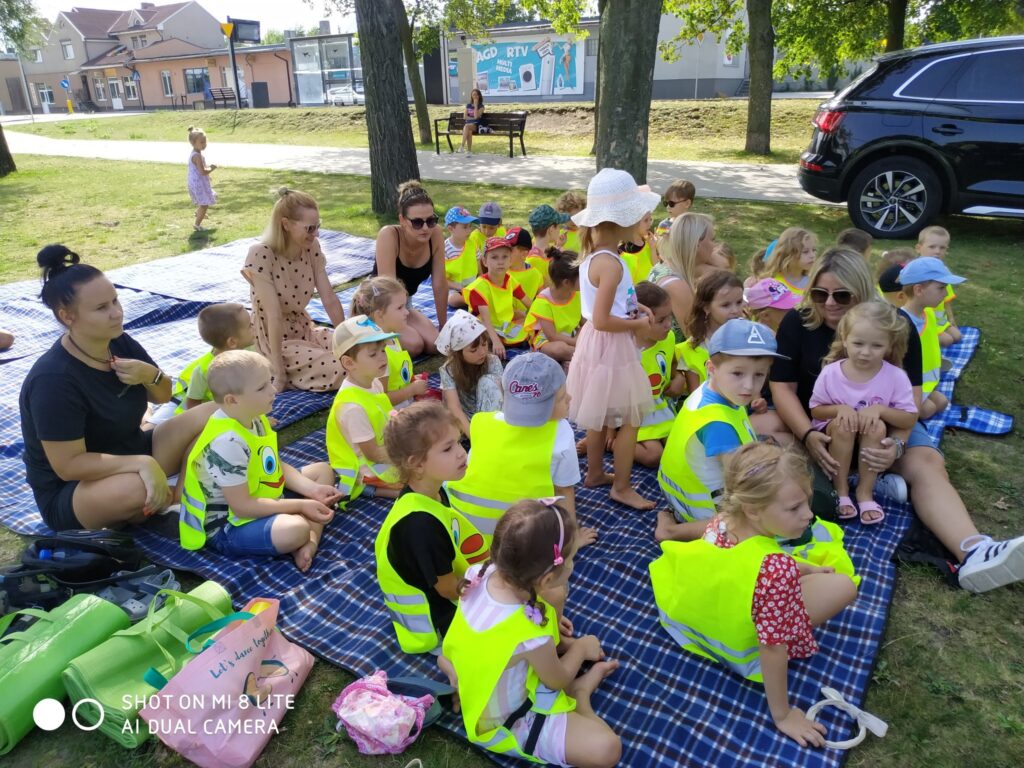 Zdjęcie przedstawia grupę przedszkolaków siedzących wraz z opiekunkami w parku na trawie. Większość dzieci ma na sobie kamizelki odblaskowe.