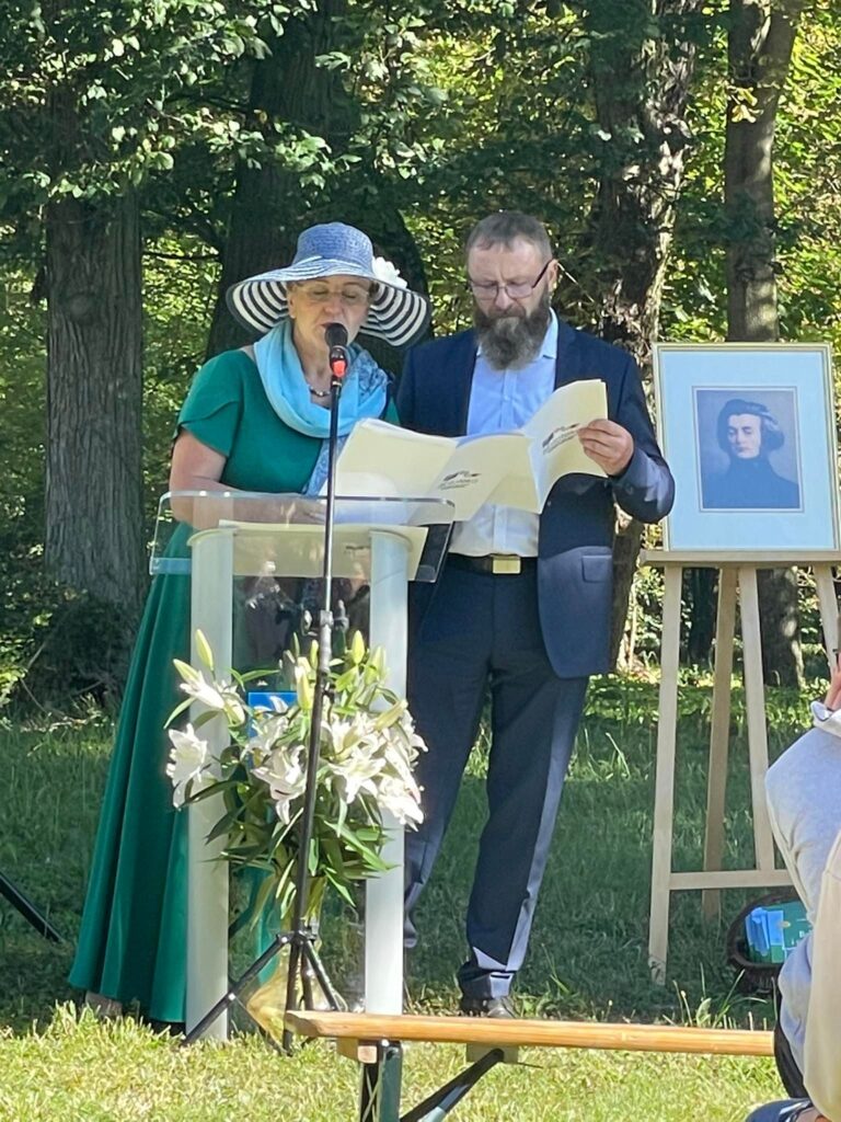 Na tle drzew parkowych pani Radna Beata Knopkiewicz wraz z mężem Pawłem, przy mównicy z mikrofonem. Obok na sztaludze portret Mickiewicza.