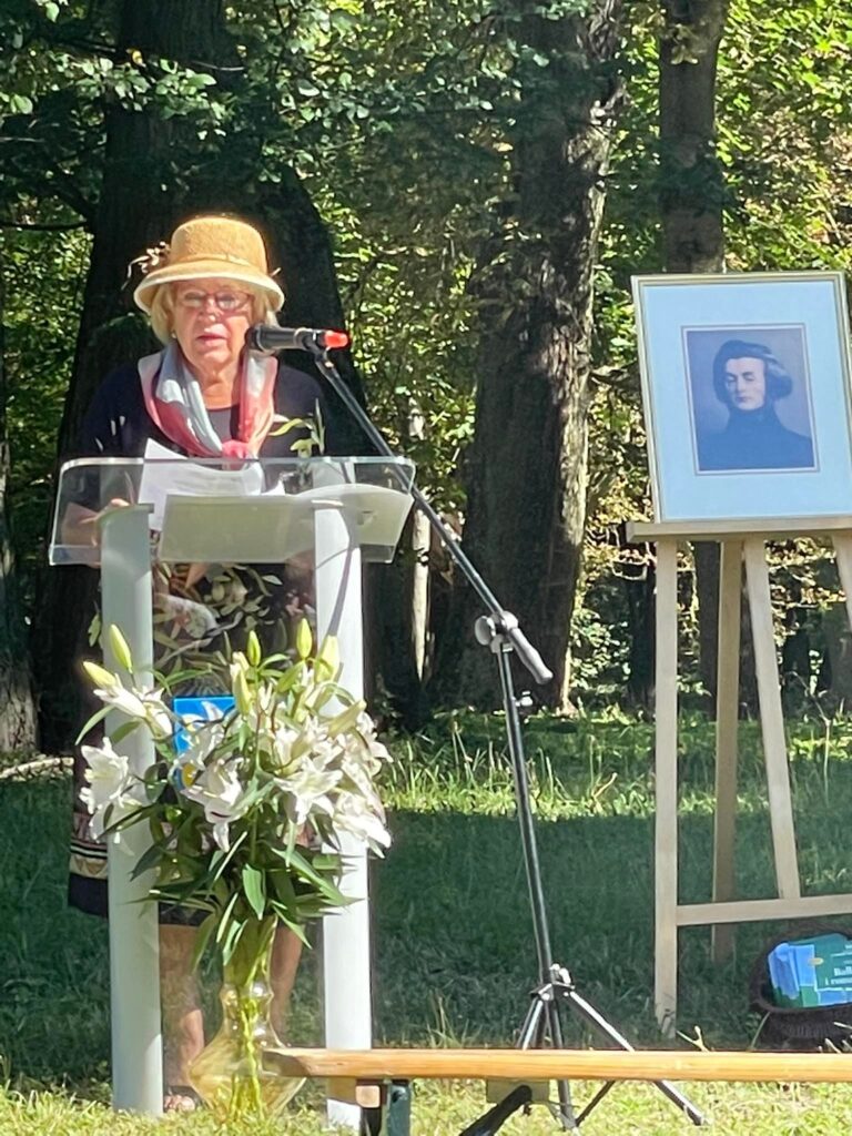 Na tle drzew parkowych pani Teresa Pokrop mównicy z mikrofonem. Obok na sztaludze portret Mickiewicza.