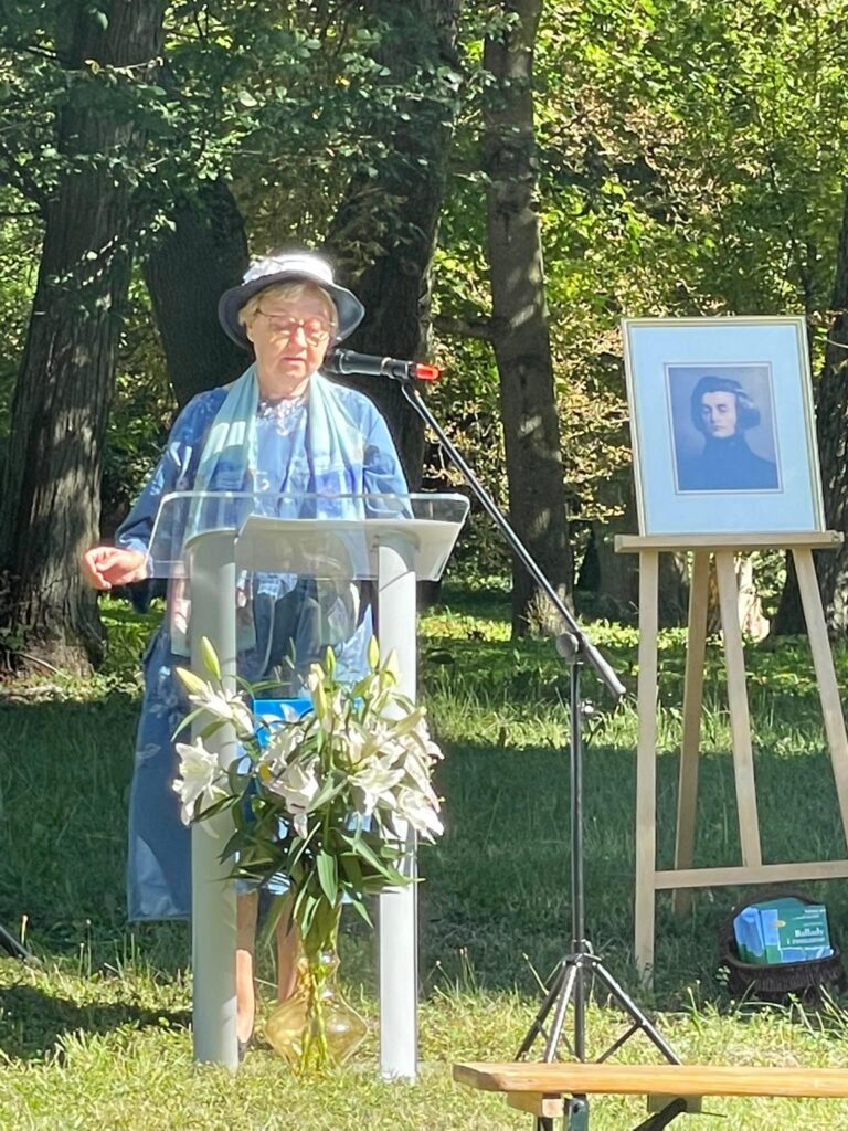 Na tle drzew parkowych pani Irena Guzińska przy mównicy z mikrofonem. Obok na sztaludze portret Mickiewicza.