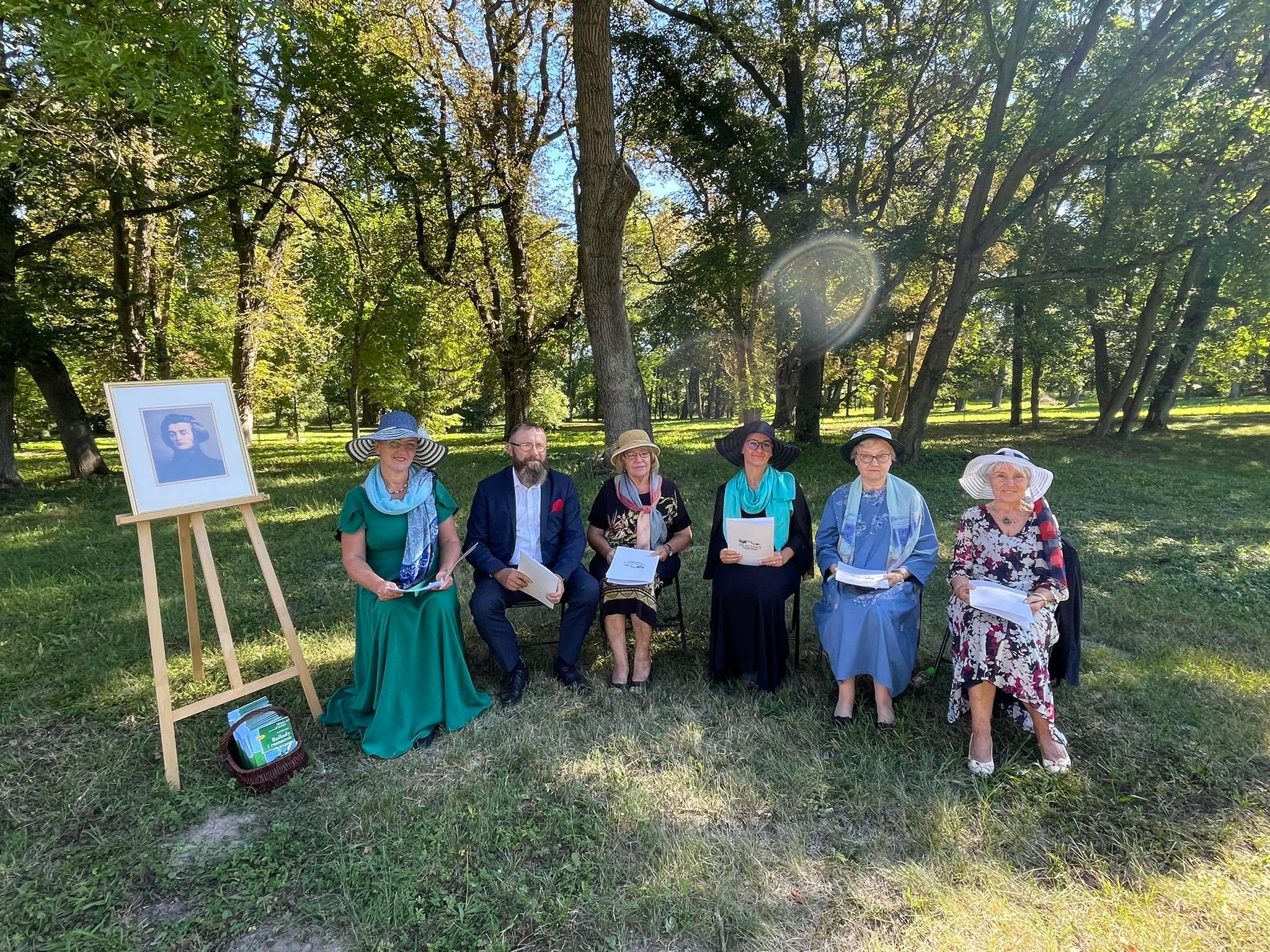 W parku, w cieniu drzew siedzi sześcioro uczestników Narodowego Czytania. Panie w kapeluszach. Po lewj stronie na sztaludze portret Mickiewicza.