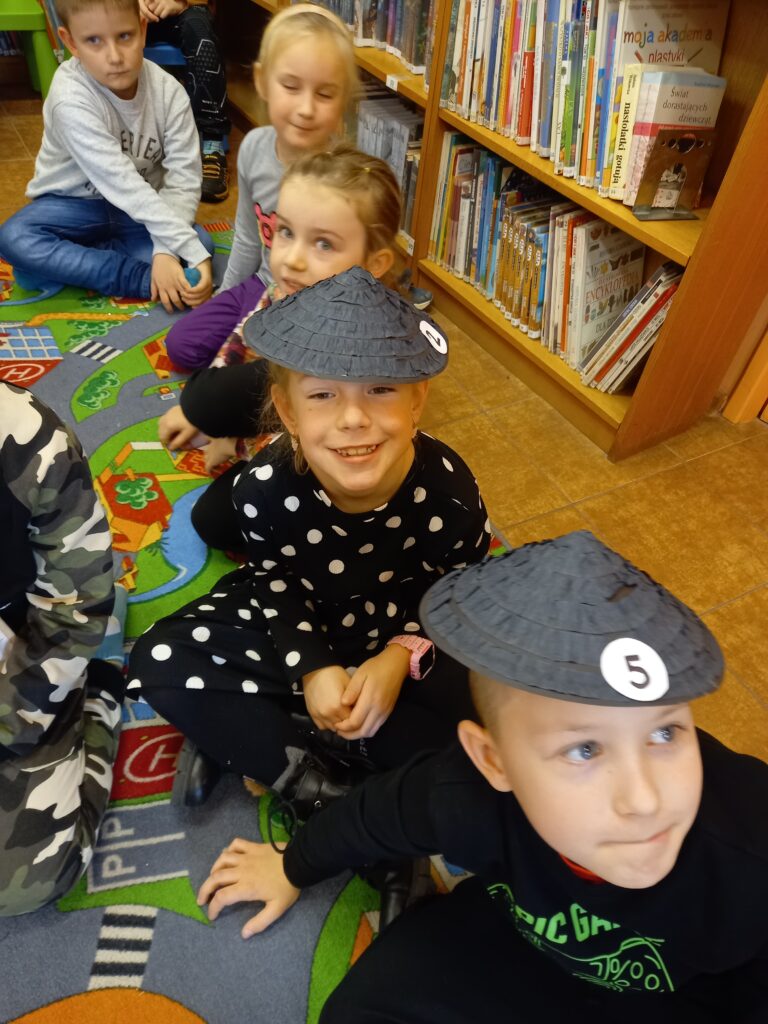 Na kolorowej wykładzinie dzieci siedzą "gęsiego". Dwoje z nich ma czarne kapelusiki.
