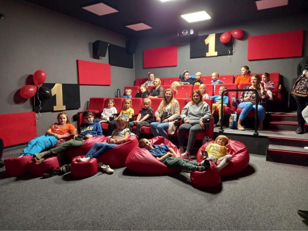 W sali kinowej o szaro-czerwonym wystroju siedzi grupa dzieci i młodzieży. Na ścianie złota jedynka.