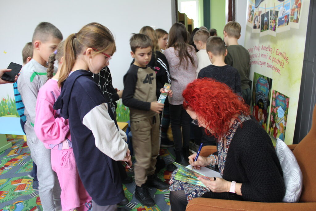 Autorka Jolanta Matuszewska siedzi w fotelu i wpisuje dedykacje do książek. Dzieci czekają w kolejce.