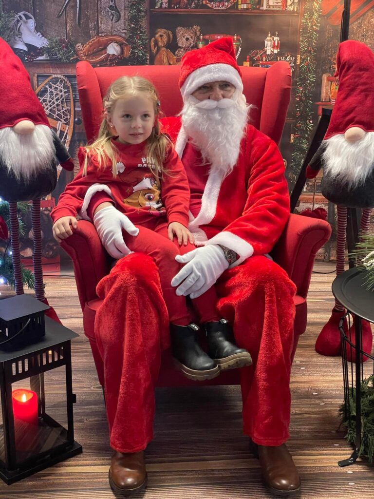 Na tle gwiazdkowych dekoracji siędzi św. Mikołaj. Na kolanach trzyma dziewczynkę.