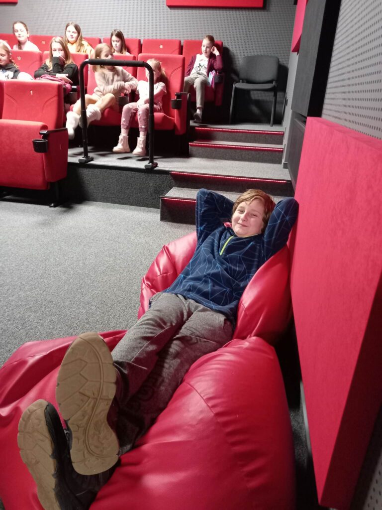 Grupa dzieci siedzi w kinie na czerwonych fotelach. Z przodu chłopiec leży na pufach.