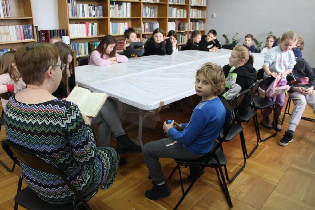 Wokół białego stołu siedzą dzieci słuchające bibliotekarki, która czyta książkę. W tle regały biblioteczne.