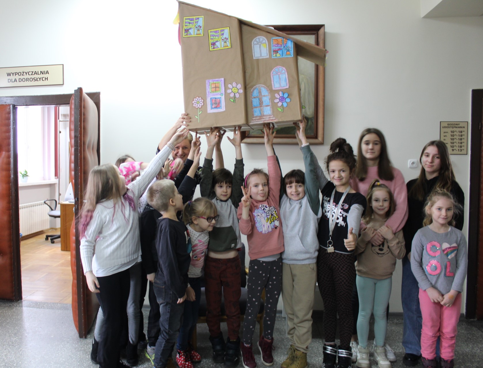Grupa dzieci w holu bibliotecznym na wysoko uniesionych rękach trzyma domek zrobiony z kartonów.