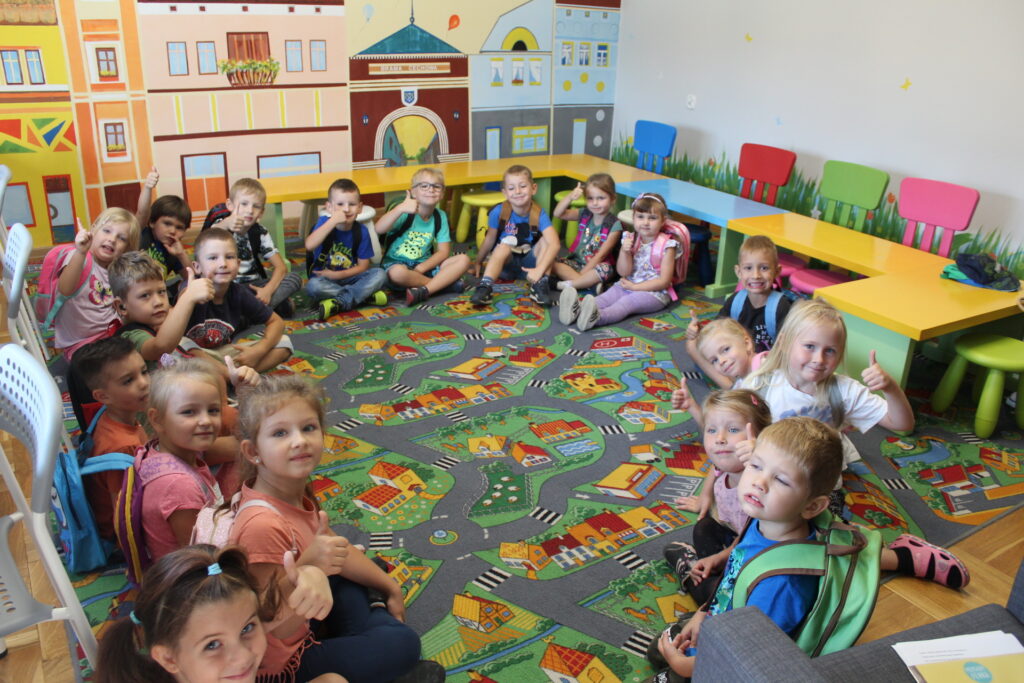 W salce bibliotecznej przedszkolaki siedzą w okręgu na dywanie.
