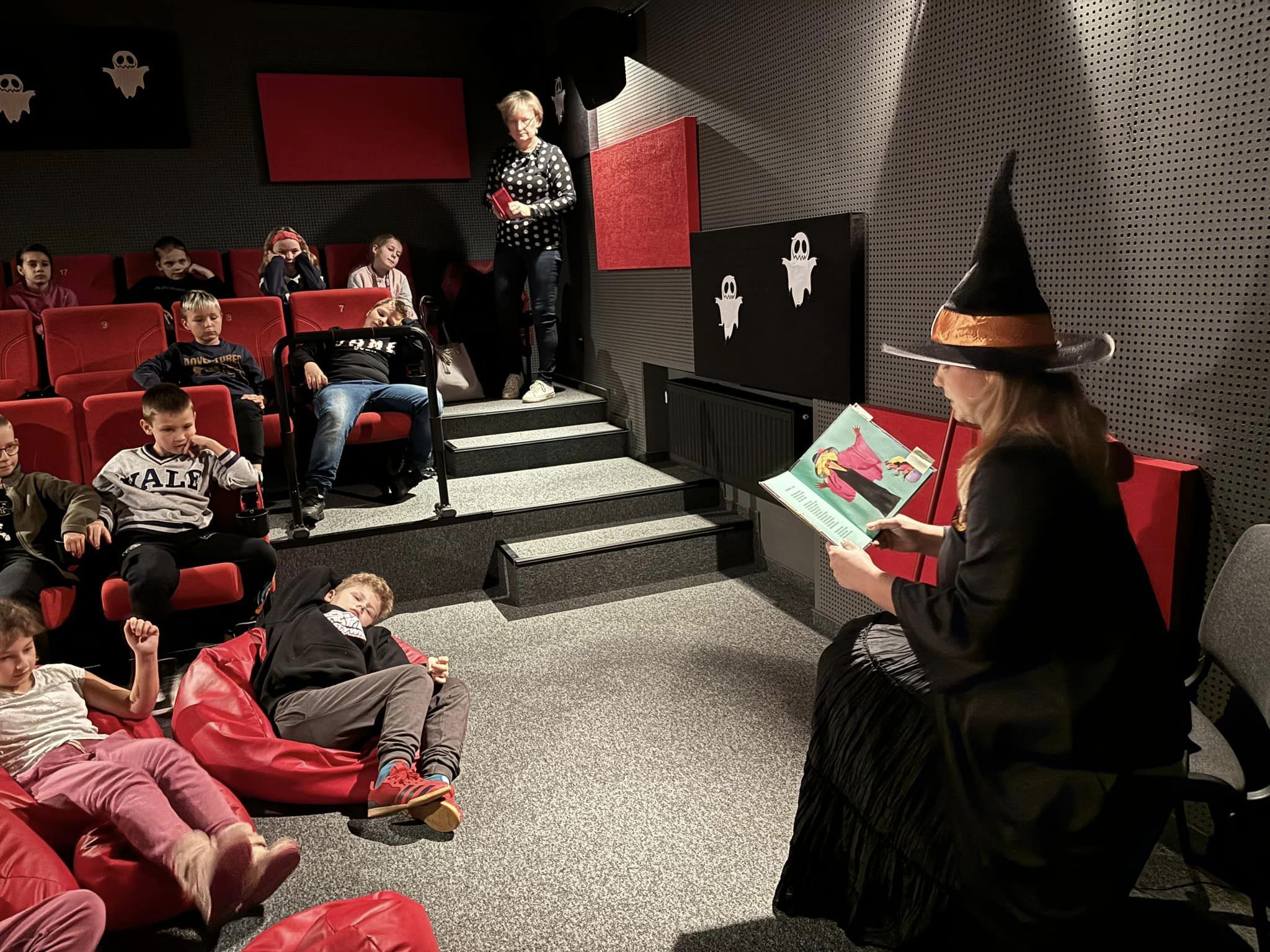 Bibliotekarka przebrana za Babę jagę czyta dzieciom bajkę w sali kinowej.