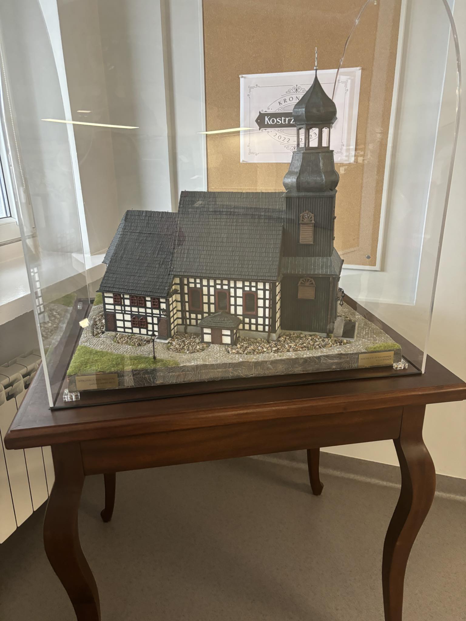 Zdjęcie przedstawia miniaturę kościoła, stojącą na drewnianym stoliku, przykrytą szklaną kopułą.
