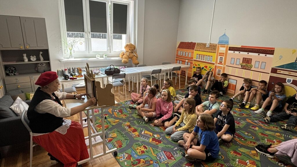 Bibliotekarka przebrana za Czerwonego Kapturka czyta siedzącym przed nią na dywanie dzieciom