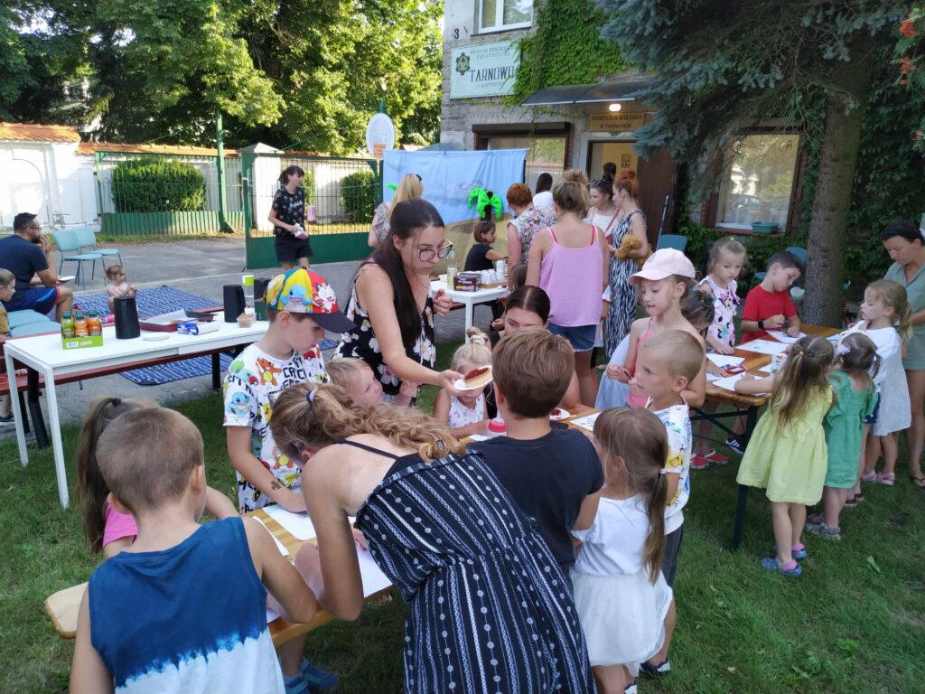 Zdjęcie przedstawia dzieci wraz z opiekunami przy rysujących przy stołach ustawionych w plenerze.