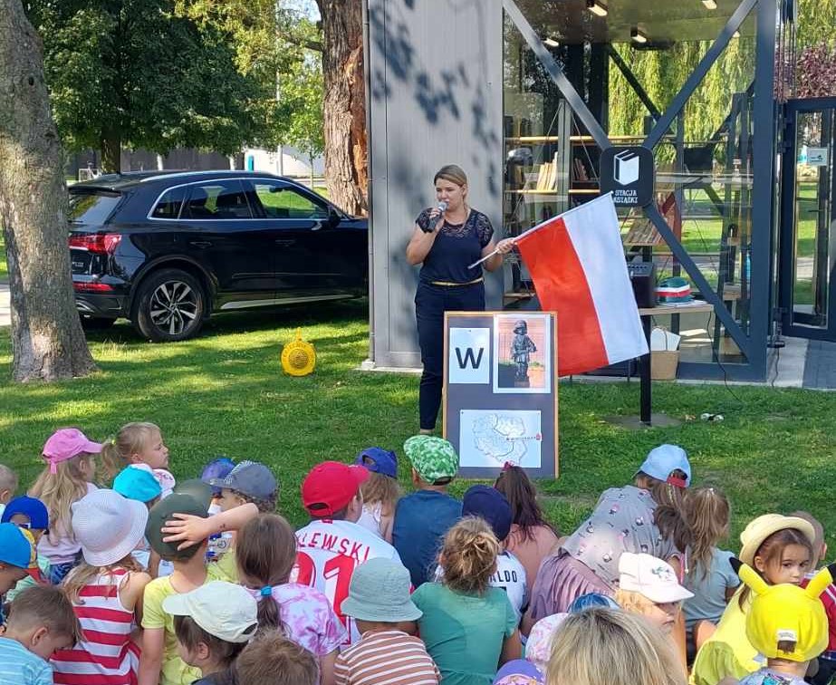 Zdjęcie przedstawia dzieci siedzące w parku przy Stacji Książki oraz prowadzącą Literacki Czwartek dyrektor biblioteki, która trzyma polską flagę.