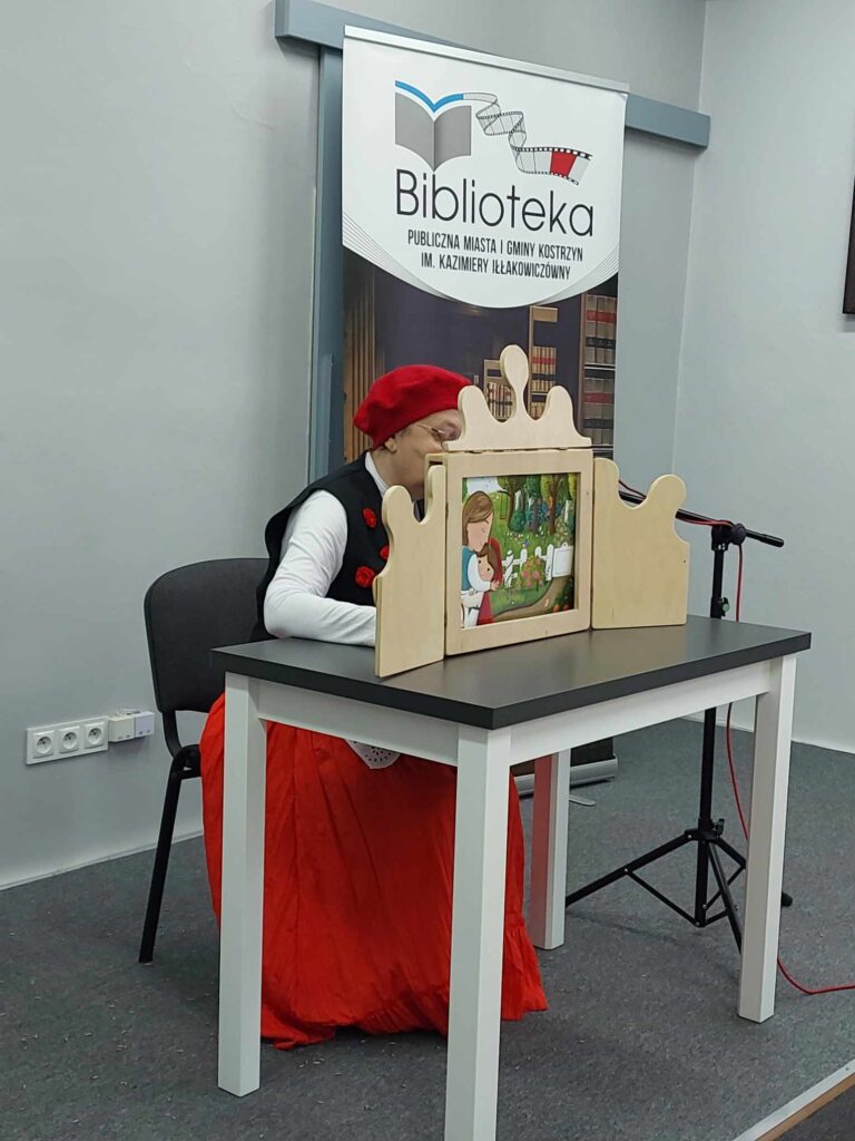 Bibliotekarka przebrana za Czerwonego Kapturka czyta bajkę. Przed nią na stoliku teatrzyk kamishibai.