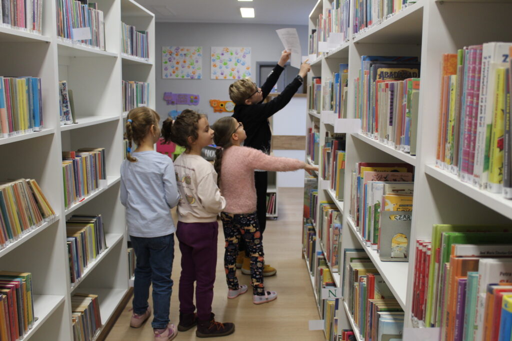 Grupa kilkunastu dzieci bierze udział z zajęciach "kodowanie na dywanie". Dzieci wyjmują książkę z bibliotecznego regału.