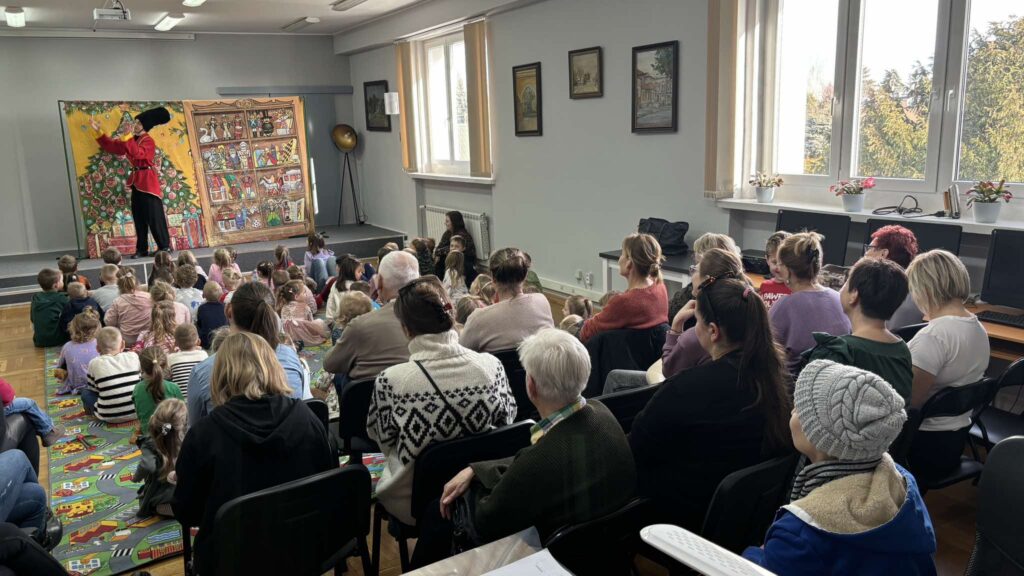 Czytelnia wypełniona dziećmi siedzącymi na dywanie. Za nimi dorośli na krzesłach. Wszyscy oglądają teatrzyk.