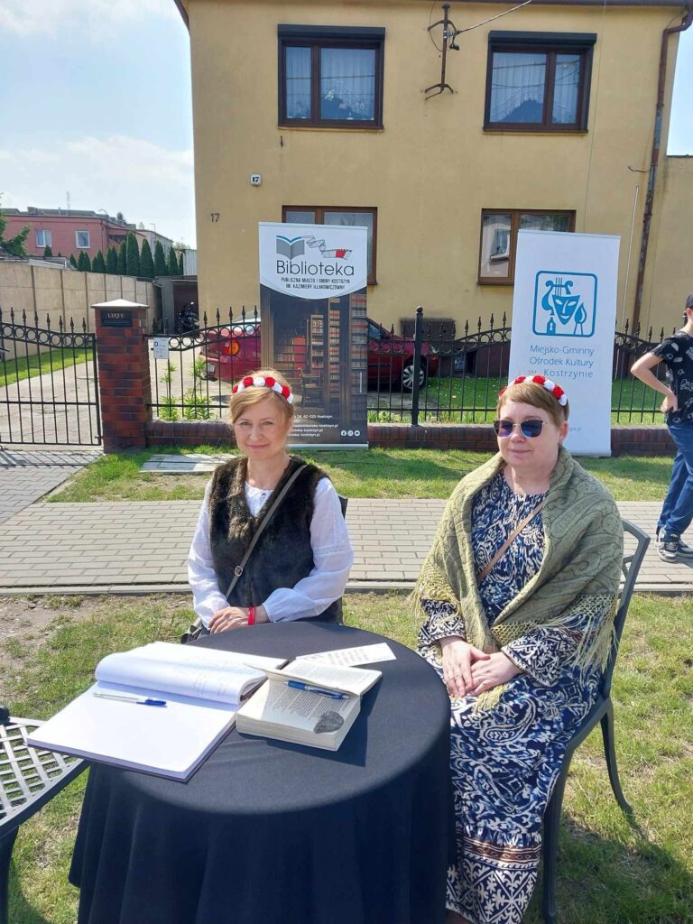 Dwie kobiety w biało-czerwonych opaskach na głowie siedzą przy stoliku w plenerze. Na stoliku widać dwie otwarte księgi