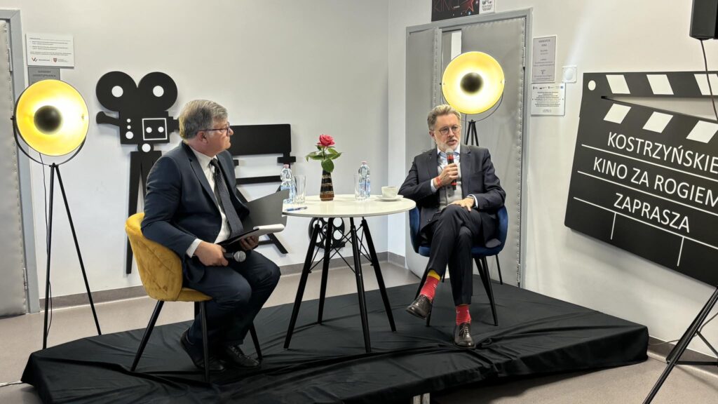 2 mężczyzn w garniturach siedzi przy stoliku na scenie i rozmawia